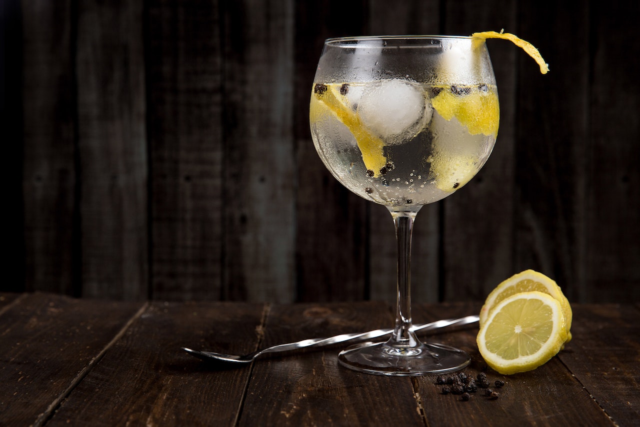 A gin cocktail with lemon peel garnish on a wooden table with a stirrer and a slice of lemon